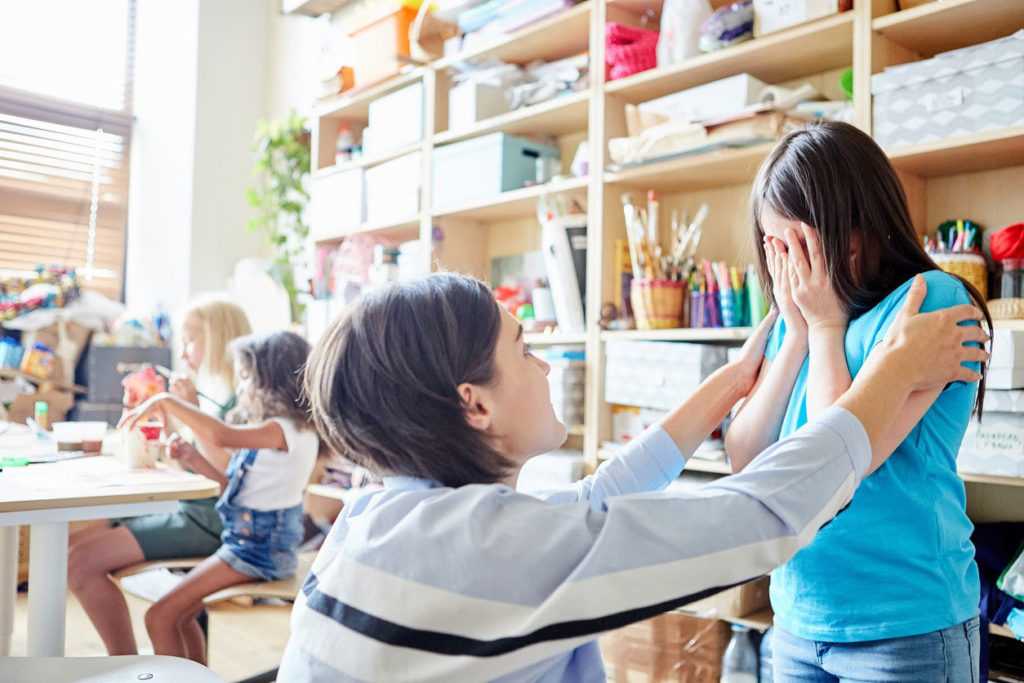 Teacher kneeling in front of a crying young girl inside a classroom