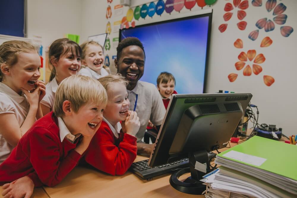 primary school students are crowded round a computer with their teacher